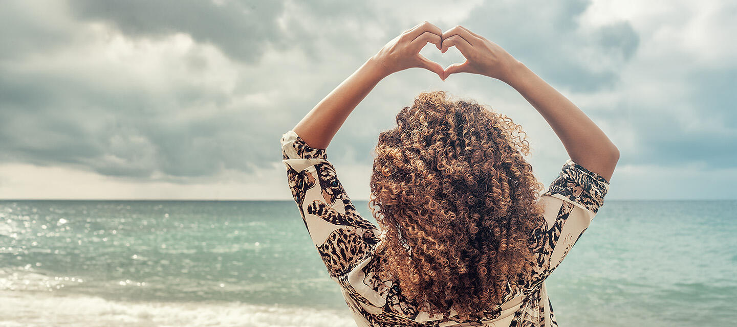 A woman with beautiful curly hair at the beach, forming a heart with her fingers above her head